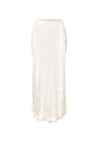 A woman wearing a white sleeveless lace-trimmed top and matching midi skirt stands outdoors against a sunlit stone wall