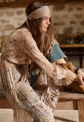 A woman in a bohemian-style outfit with a paisley-print long-sleeve top and matching headband, leaning on a rustic wooden table in a warm, indoor setting.