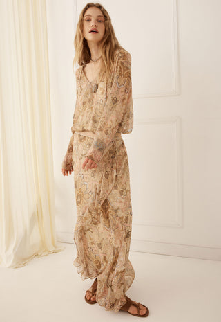 A woman modeling a long-sleeved maxi dress with a beige and brown paisley pattern, standing in a bright, minimalist room with white walls and a cream curtain.