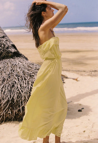 A woman wearing a strapless pale yellow maxi dress stands on a sandy beach with the ocean and a thatched palm tree in the background.