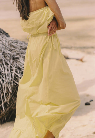 A woman wearing a flowing pale yellow strapless maxi dress standing on a sandy beach next to a thatched structure.