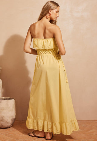 A woman wearing a strapless yellow maxi dress with ruffled trim and a tie waist, shown from the back in a warm-toned indoor setting.