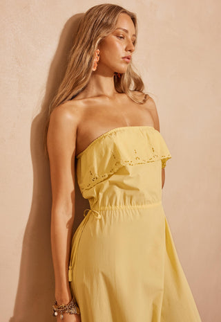 A woman wearing a strapless pale yellow dress with eyelet lace detailing on the bodice, standing against a textured beige wall.