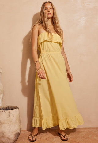 A woman modeling a strapless yellow maxi dress with ruffle details, standing against a textured beige wall.