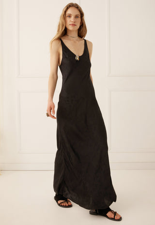 A woman modeling a black sleeveless maxi dress with a deep V-neckline and a small silver pendant at the neckline, standing against a white paneled wall.