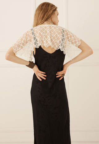 A woman shown from the back wearing a black maxi dress with a sheer white lace cape-style shoulder overlay, standing against a white paneled wall.