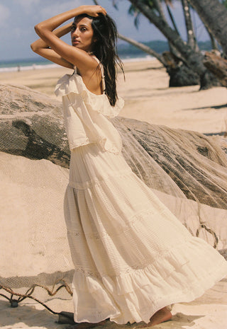 A woman wearing a flowing cream boho-style maxi dress stands on a sandy beach next to a large driftwood log