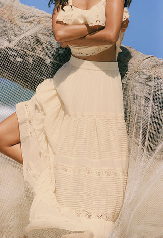 A woman wearing a cream-colored tiered maxi dress with delicate lace and embroidery details, posed outdoors against a blue sky with sheer netting draped around her.