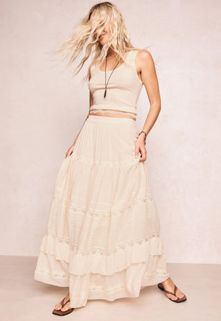 A woman modeling a cream-colored two-piece outfit consisting of a sleeveless crop top and a long tiered maxi skirt with delicate lace trim, paired with brown flip-flop sandals against a textured beige studio backdrop.