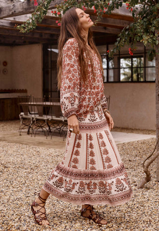 A woman wearing a bohemian-style two-piece dress set with a rust brown and cream floral pattern, walking outdoors on a gravel path near a patio.